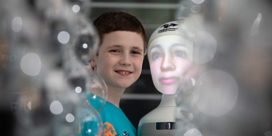 Young boy with Furhat robot head, standing behind some glass bubbles