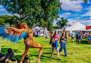 http://two%20people%20walking%20through%20a%20music%20festival%20in%20the%20sunshine.%20On%20the%20left%20is%20a%20person%20dressed%20as%20an%20angel,%20posing%20with%20their%20arms%20and%20head%20stretched%20backwards
