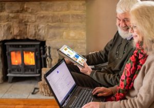 http://Older%20man%20and%20woman%20using%20a%20tablet,%20iPad%20and%20laptop%20by%20a%20lit%20stove.