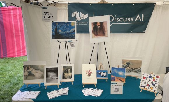 A table with a teal green tablecloth. On the table there are a number of artworks displayed on easels. Behind the table, there is a banner with the ADAPT logo and the #DiscussAI logo.