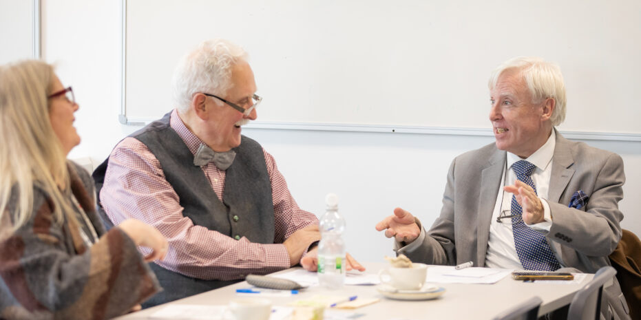 Photo of two older gentlemen sitting at a table in a classroom-like room, smiling and deep in conversation
