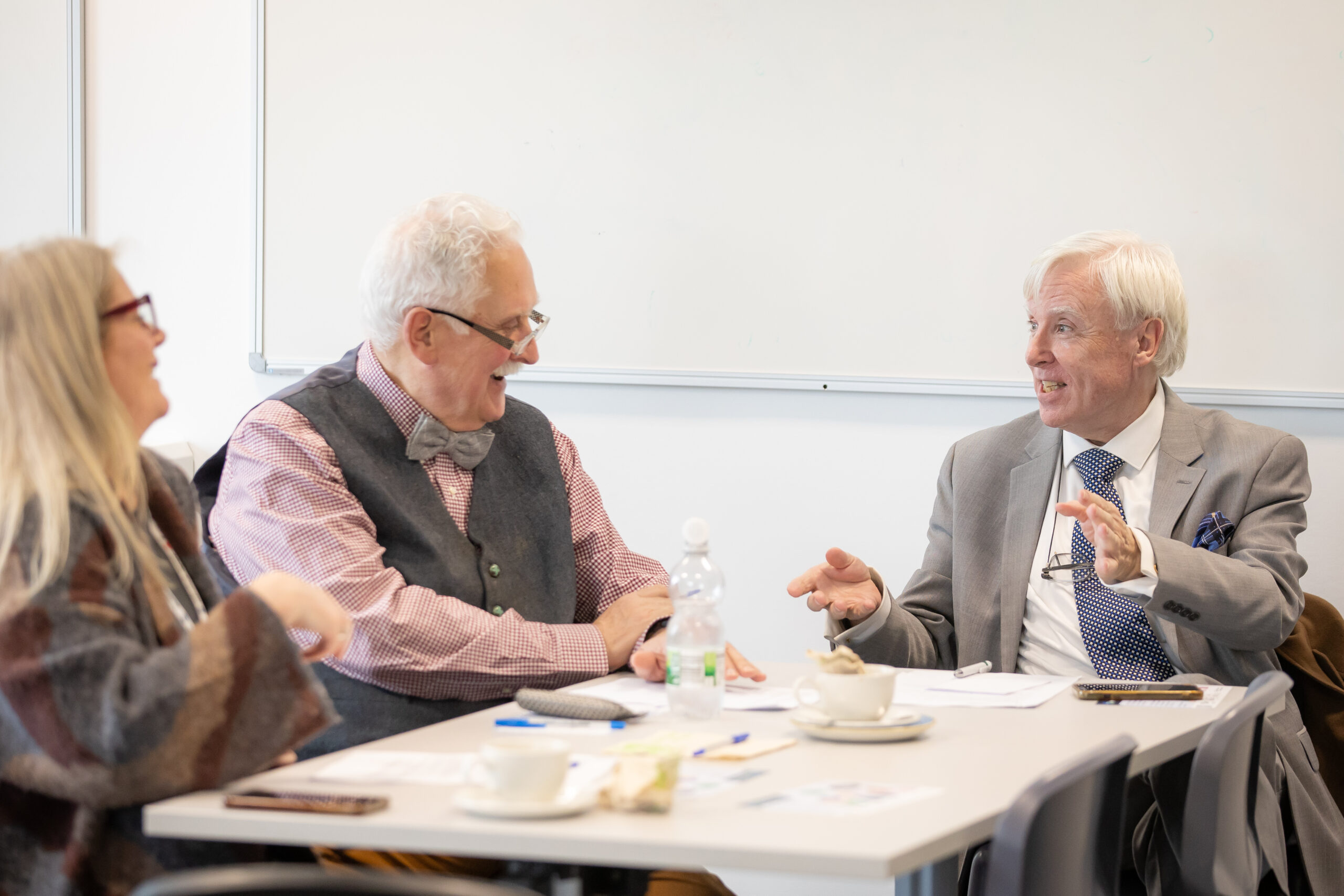 Photo of two older gentlemen sitting at a table in a classroom-like room, smiling and deep in conversation