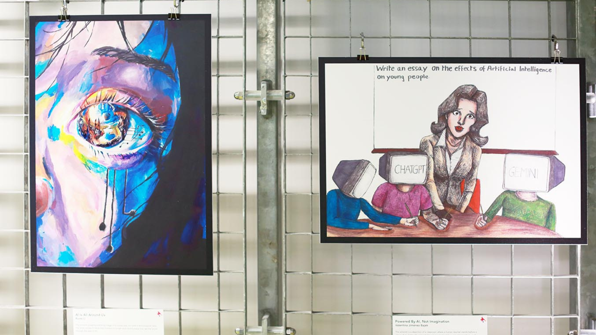 A pair of art pieces are displayed on a gray wire mesh wall, possibly at a school or public exhibition. The piece on the left is a vibrant, close-up portrait of an eye and the surrounding part of a face. The style is abstract and expressive, using a mix of dark and bright colors like deep blues, purples, pinks, and yellows. The eye itself seems to be mechanical or digital, with an intricate iris made of what look like circuits and gears. The person's face appears to be crying, with a streak of blue and white tears running down the cheek. A small, white label with text is visible below the artwork. The piece on the right is a satirical or critical cartoon. It shows a woman, possibly a teacher, sitting at a desk with a concerned expression. She is looking at two figures in front of her, both with television-like screens for heads. The screen on the left says "CHATGPT" and the screen on the right says "GEMINI." A thought bubble above the woman's head contains the text, "Write an essay on the effects of Artificial Intelligence on young people." This piece also has a label below it.