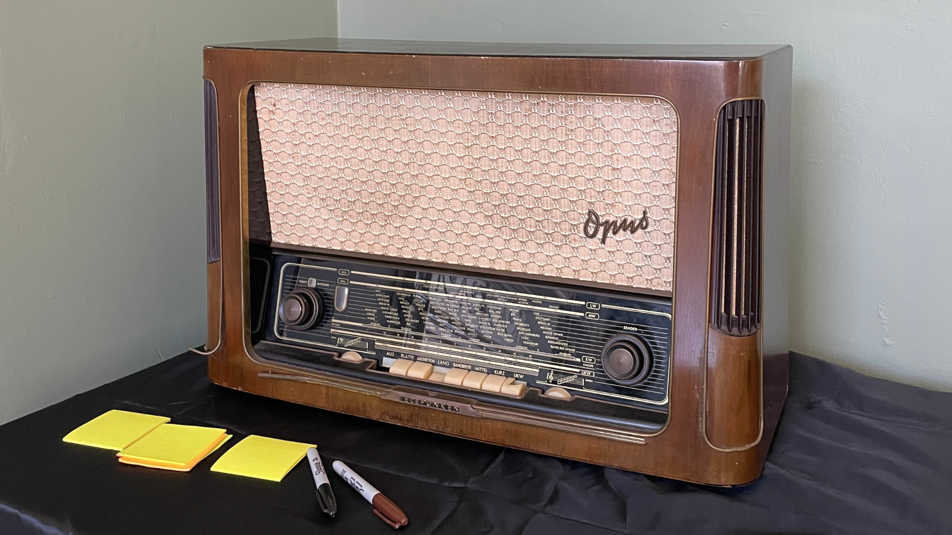 A front-on view of a large, vintage tube radio. It has a rich, dark wooden cabinet with a rectangular shape and rounded edges. The front features a large, off-white mesh speaker grille with a quilted texture. The word "Opus" is written in cursive script on the top right of the grille. Below the grille is a tuning dial with various frequencies and station names, flanked by two large, circular knobs. A row of smaller push buttons sits along the bottom edge of the dial. The radio is sitting on a black cloth, with a stack of yellow sticky notes and two markers in the foreground. The background is a plain, light-green wall.