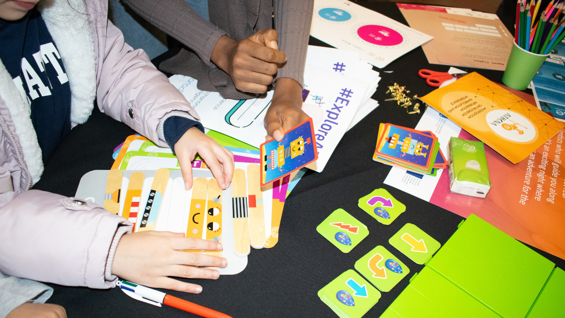 A top-down view of two people's hands and arms at a table, engaged in a hands-on activity. A person on the left is wearing a light purple puffy jacket over a dark blue shirt and is holding and sorting several laminated cards. The cards have a series of cute, yellow robotic characters on them. The person on the right, wearing a gray long-sleeved shirt, is holding up a single card with a similar robot character on it. Spread out on the black tabletop are various items for the activity. There are several small, bright green square tiles with different arrows and symbols on them. Some paper instructions are also visible, one with the hashtag #explore. A pair of scissors, a green cup with colored pencils, and an orange document are also on the table.