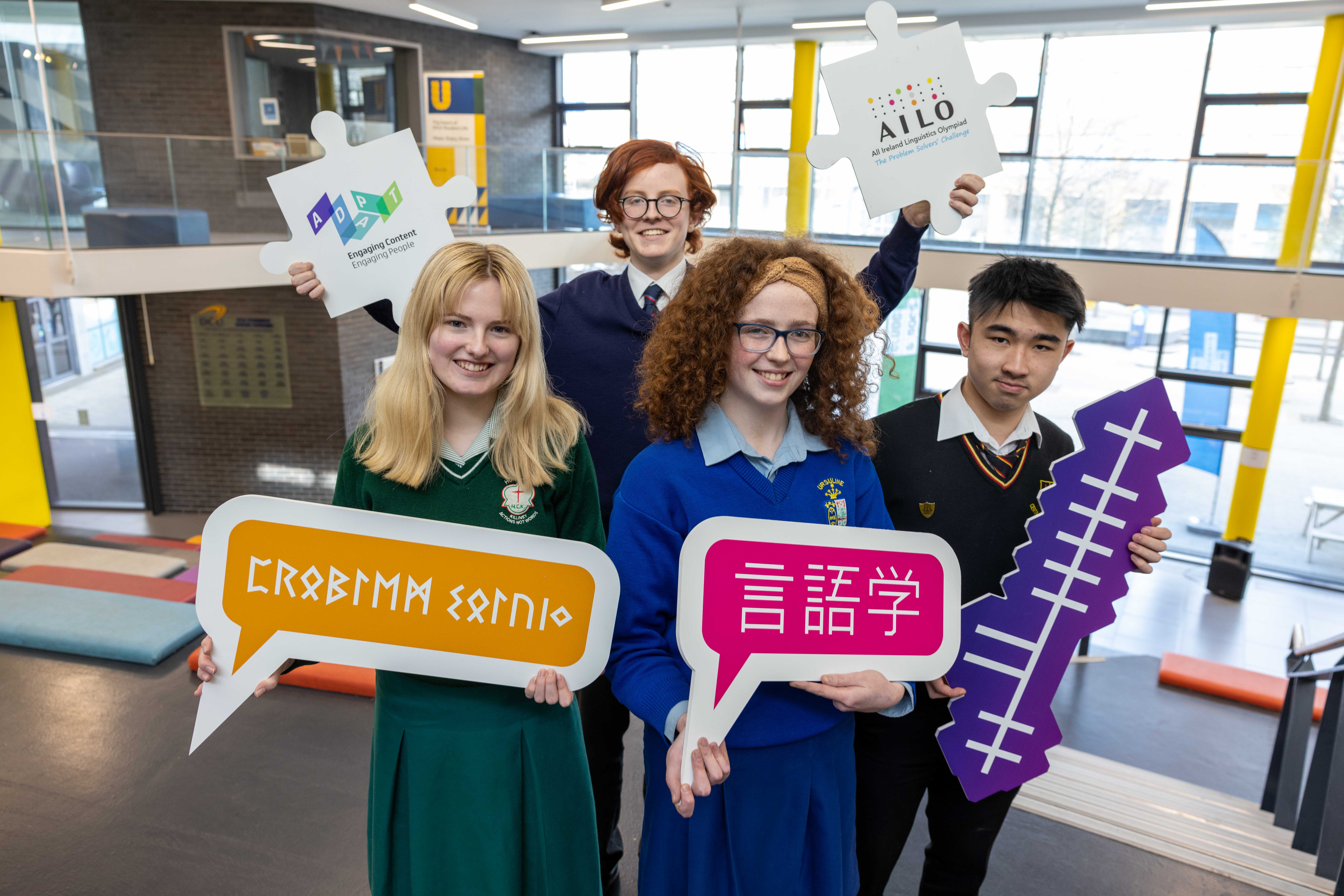 Four smiling teenage students in school uniforms hold up cut-out signs depicting foreign language scripts. One student holds jigsaw-shaped signs with the ADAPT and AILO logos.