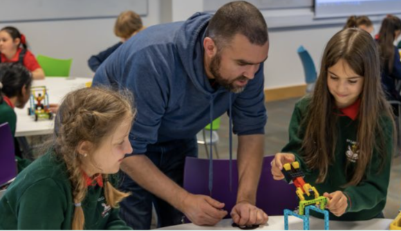 Photo of a male teacher helping two young female students in school uniforms with a Lego-like game