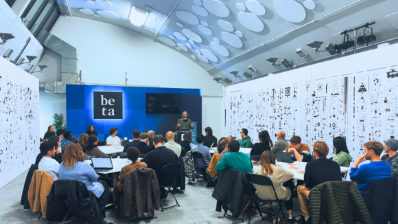 Photo from the AI Art Assembly 2024 with adults sitting around circular tables paying attention to a speaker at a lectern with the sign "Beta" behind him