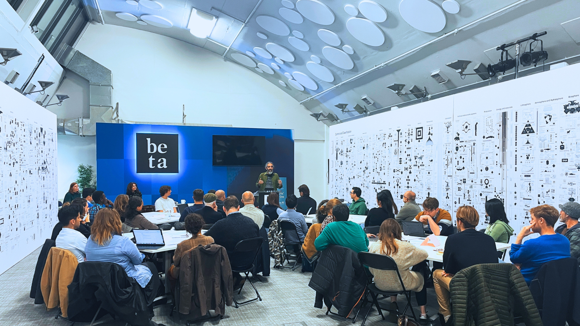 Photo from the AI Art Assembly 2024 with adults sitting around circular tables paying attention to a speaker at a lectern with the sign "Beta" behind him